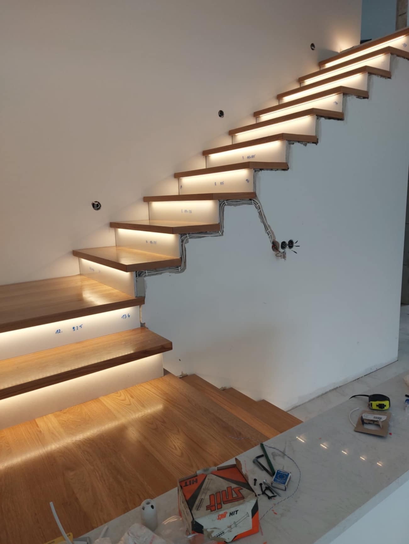 Drewniane schody z podświetleniem LED w trakcie montażu, widoczne przewody elektryczne i narzędzia na blacie obok.
