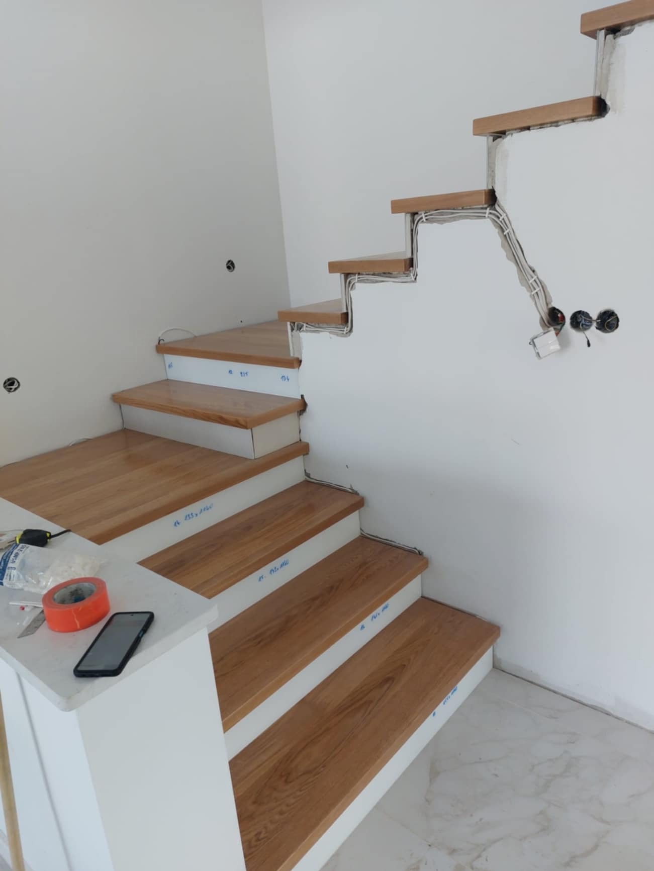 Schody drewniane w trakcie montażu, widoczne przewody elektryczne i narzędzia na blacie, częściowo wykończone.