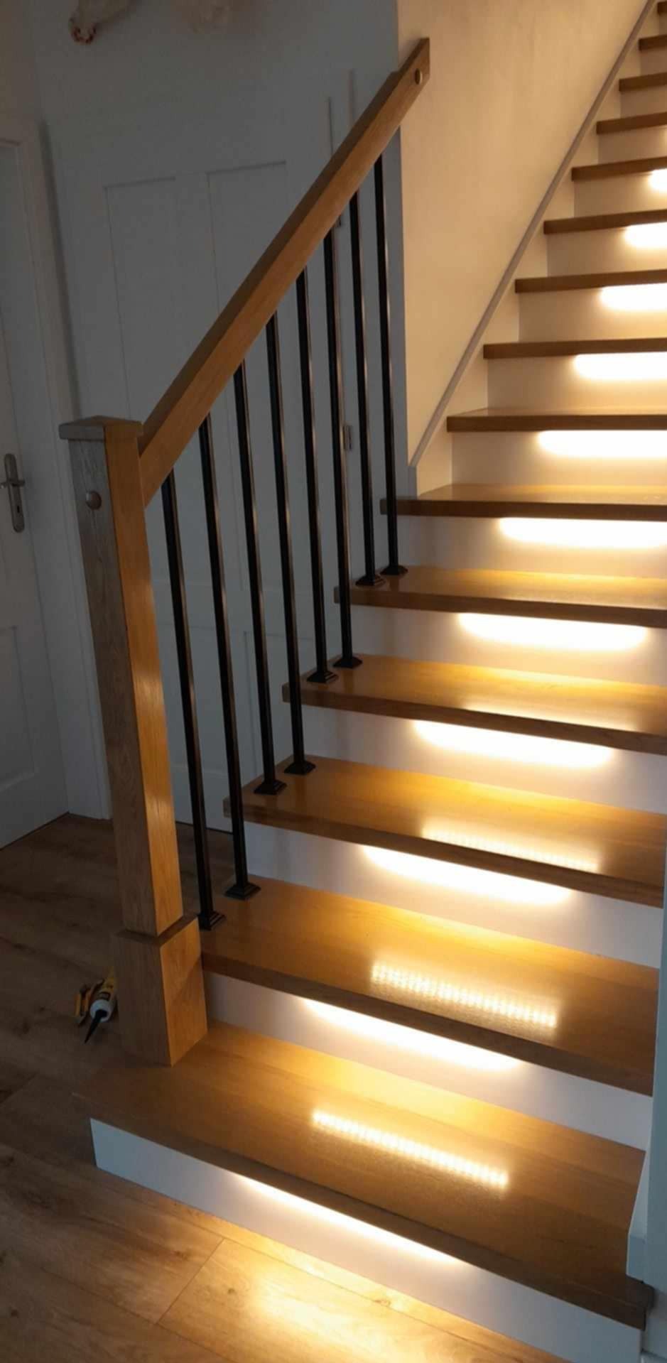 Drewniane schody z podświetleniem LED pod każdym stopniem, czarne metalowe balustrady, widoczny fragment podłogi i drzwi, perspektywa od dołu.