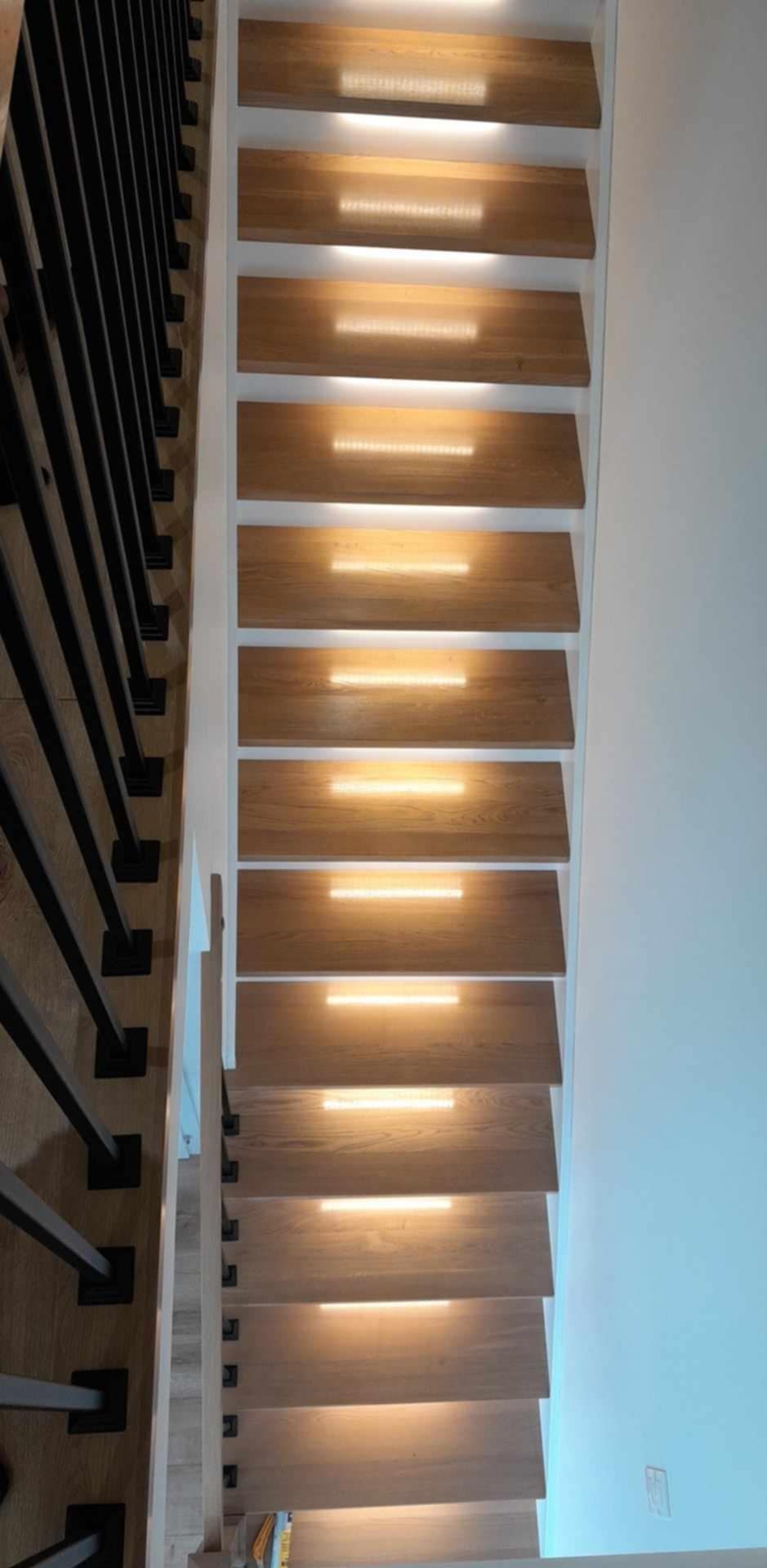 Drewniane schody z wbudowanym oświetleniem LED, czarne metalowe balustrady po lewej stronie, białe ściany.
