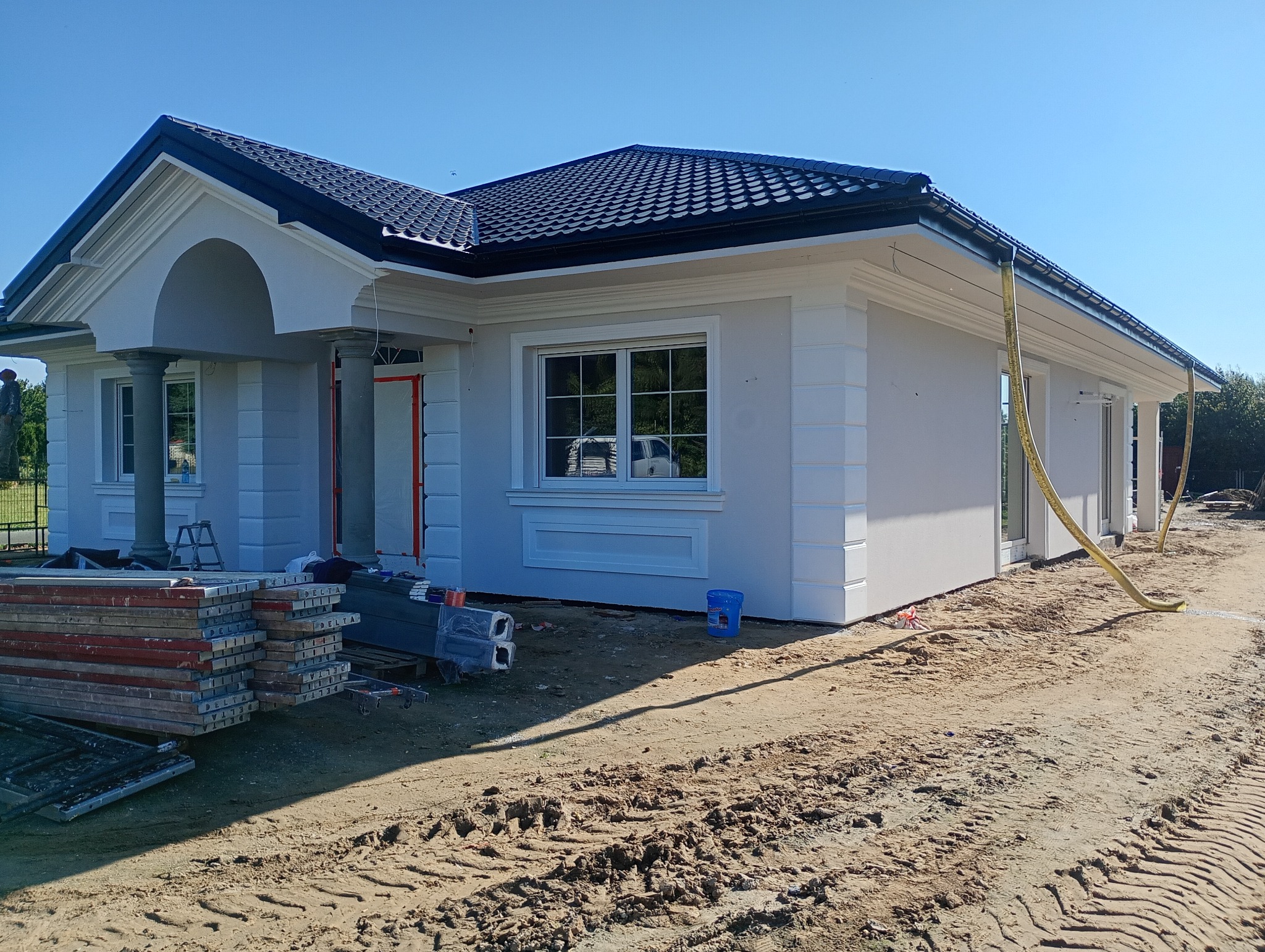 Jasny dom jednorodzinny z ciemną dachówką, widoczne elementy dekoracyjne elewacji, teren wokół w trakcie prac wykończeniowych, błękitne niebo.