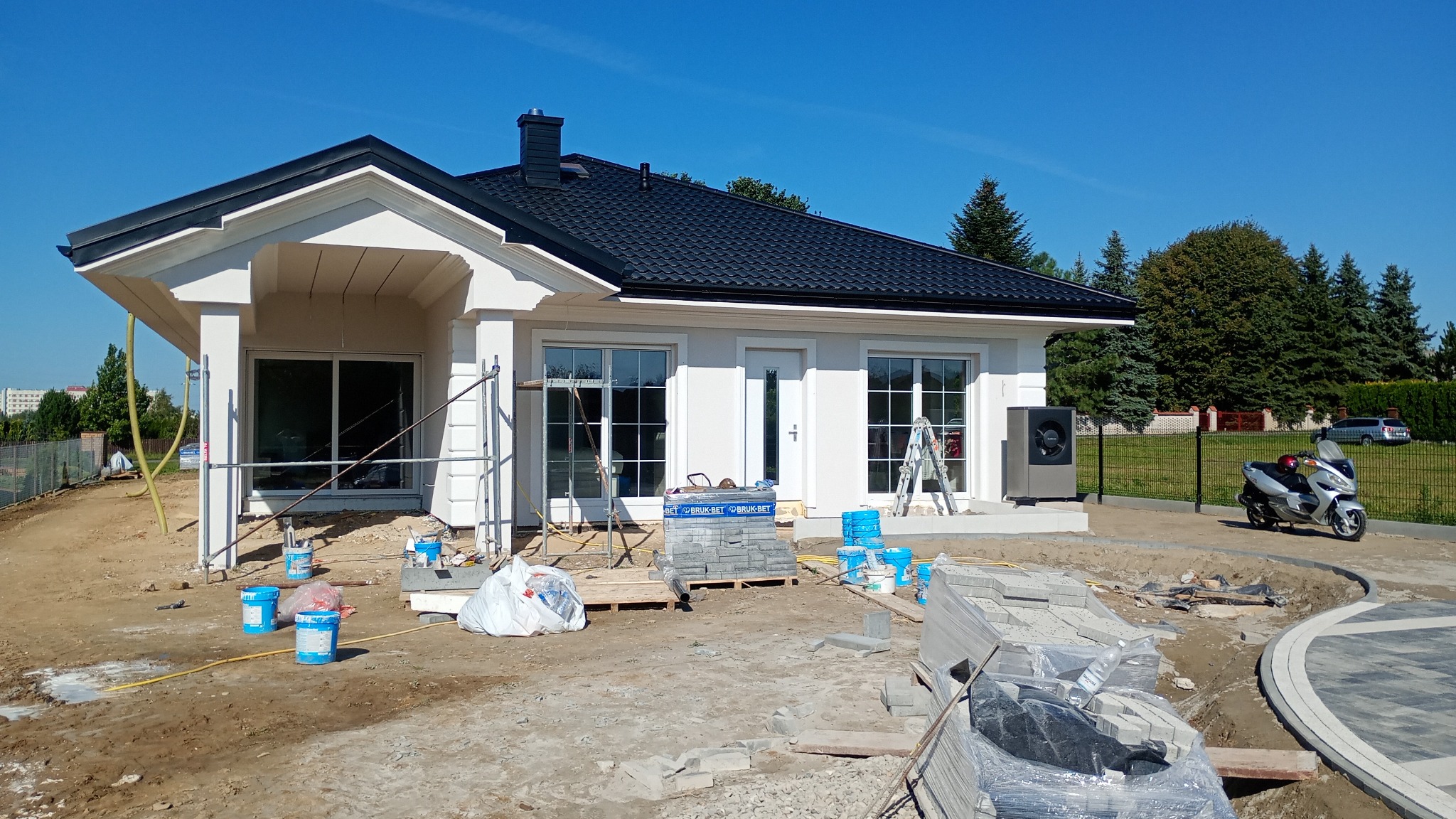Jasny dom jednorodzinny z czarnym dachem w trakcie prac wykończeniowych elewacji zewnętrznej. Widoczne rusztowania, materiały budowlane i skuter.