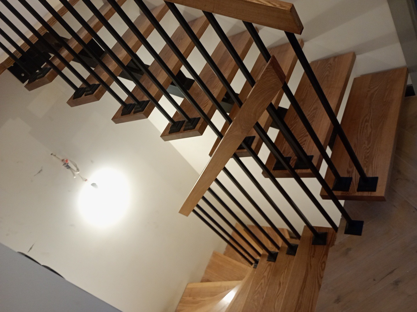 Drewniane schody z czarnymi, metalowymi balustradami, widziane z góry pod kątem. Jasne drewno stopni kontrastuje z ciemnymi elementami konstrukcyjnymi.