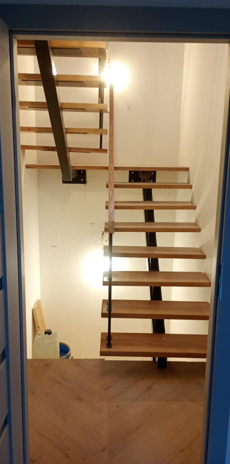 Nowoczesne schody drewniane z metalowymi elementami, widoczne z dołu. Podłoga wyłożona parkietem w jodełkę. W tle białe ściany i widoczne narzędzia.