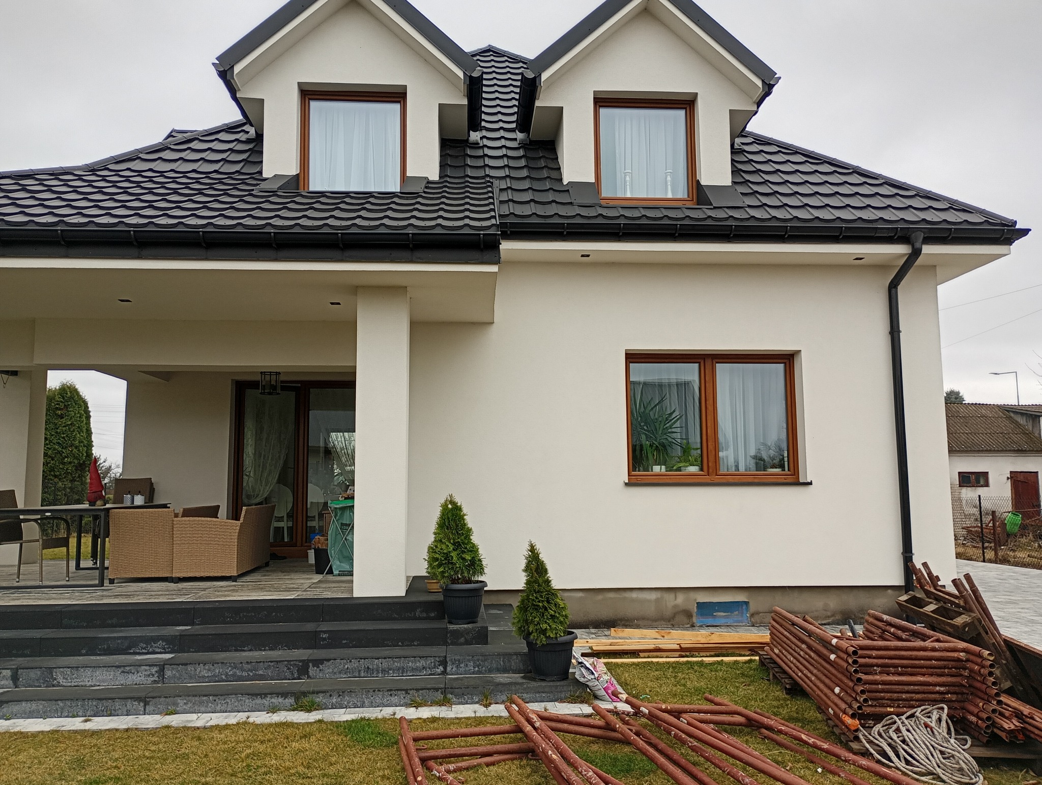 Elewacja domu jednorodzinnego z czarną dachówką i białymi ścianami. Widoczne okna z brązowymi ramami, elementy rusztowania i rośliny ozdobne.