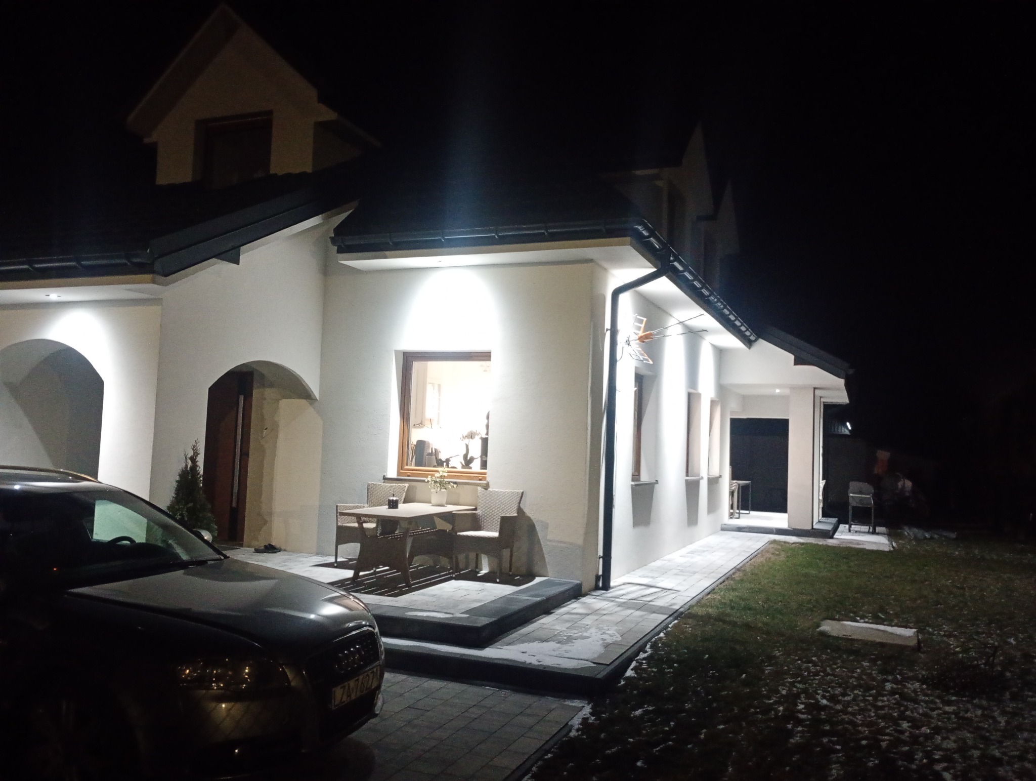 Oświetlona elewacja domu jednorodzinnego nocą, z widocznym fragmentem samochodu i meblami ogrodowymi na tarasie. Białe ściany, ciemny dach, nowoczesny styl.