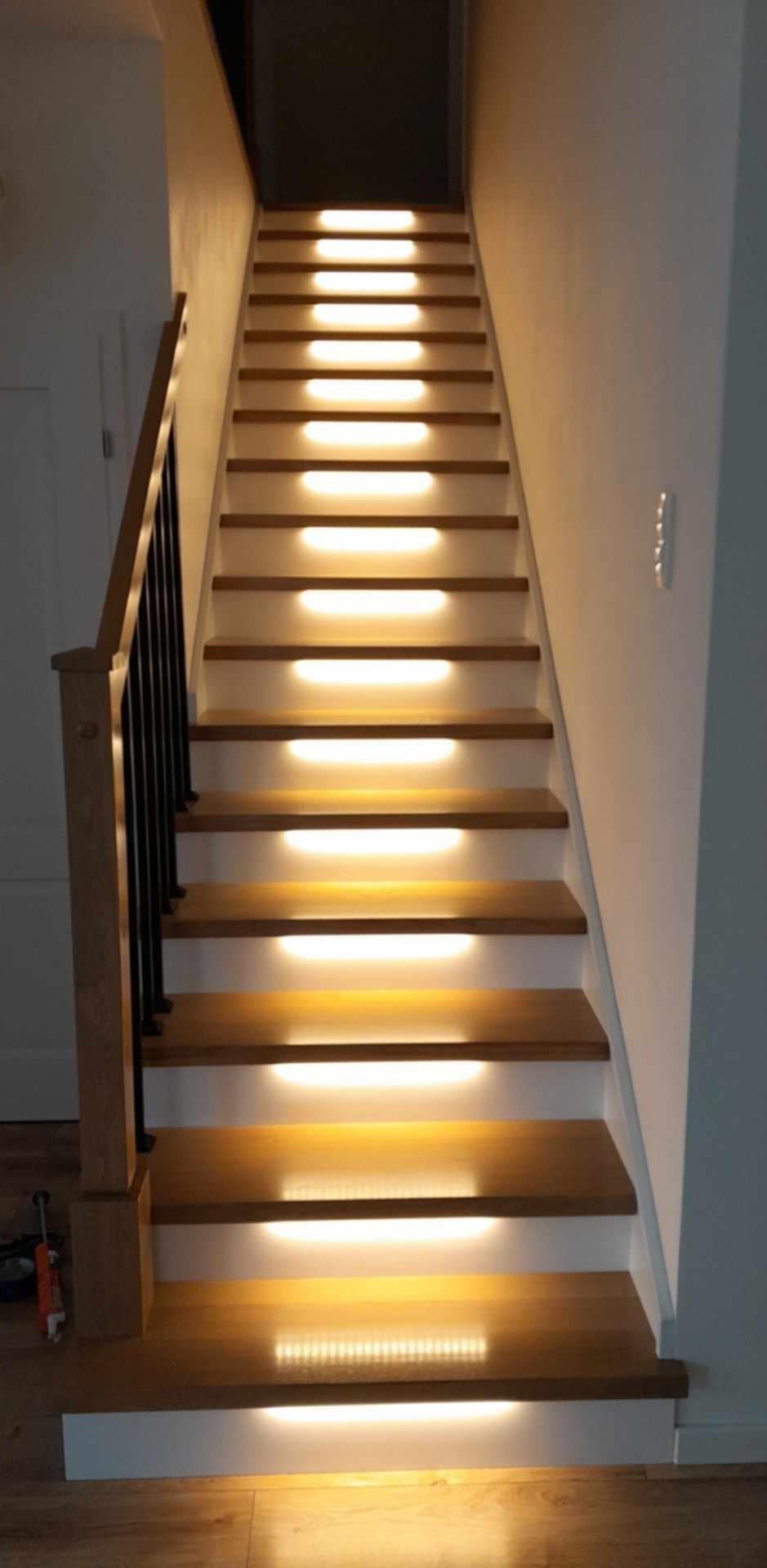 Drewniane schody z podświetleniem LED, czarna balustrada. Ciepłe światło podkreśla naturalny odcień drewna. Widok z dołu na górę, nowoczesny design.