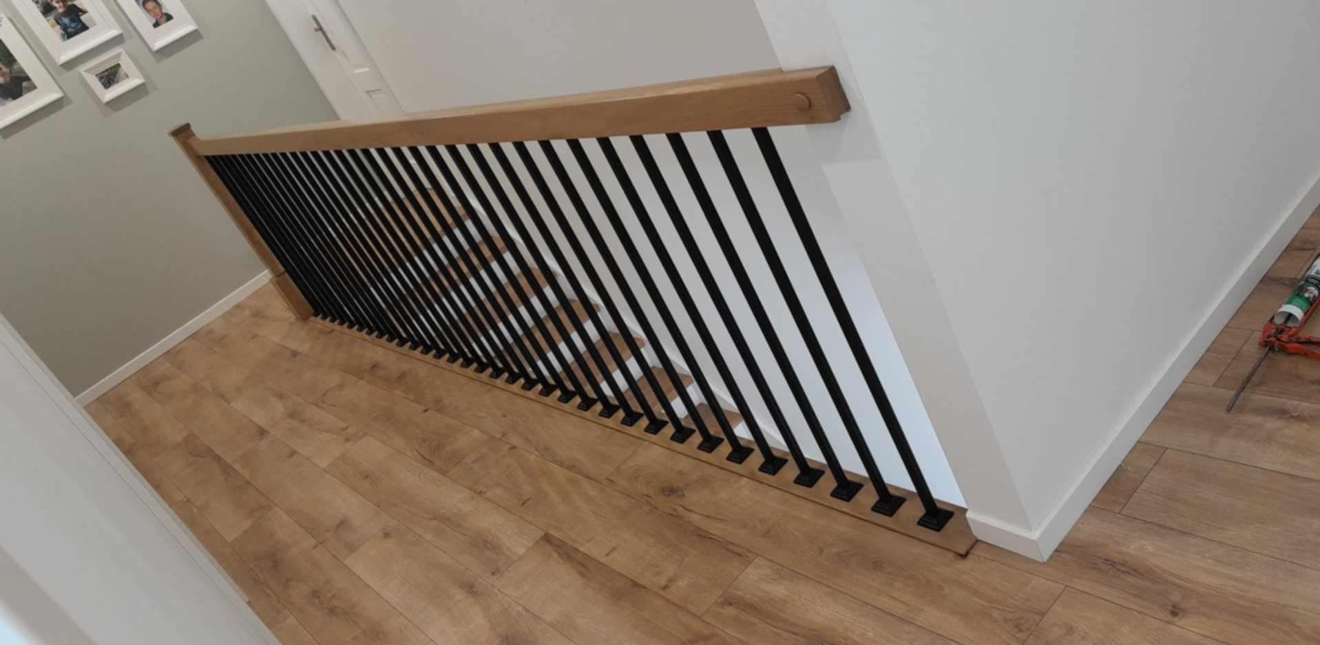 Nowoczesne schody z drewnianą poręczą i czarnymi, pionowymi balustradami, widok z góry na parkietową podłogę. Minimalistyczny design, jasne ściany, wykończenie wnętrza.