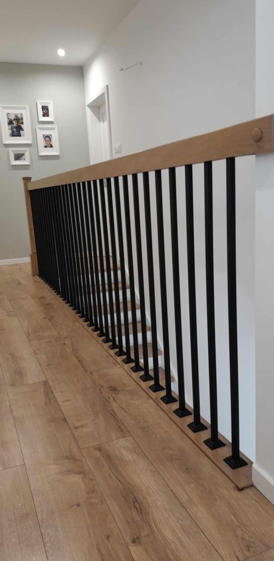 Nowoczesna balustrada schodowa z czarnymi, pionowymi szczeblami i drewnianym poręczą, widoczna na tle jasnych ścian i drewnianej podłogi. Minimalistyczny design.