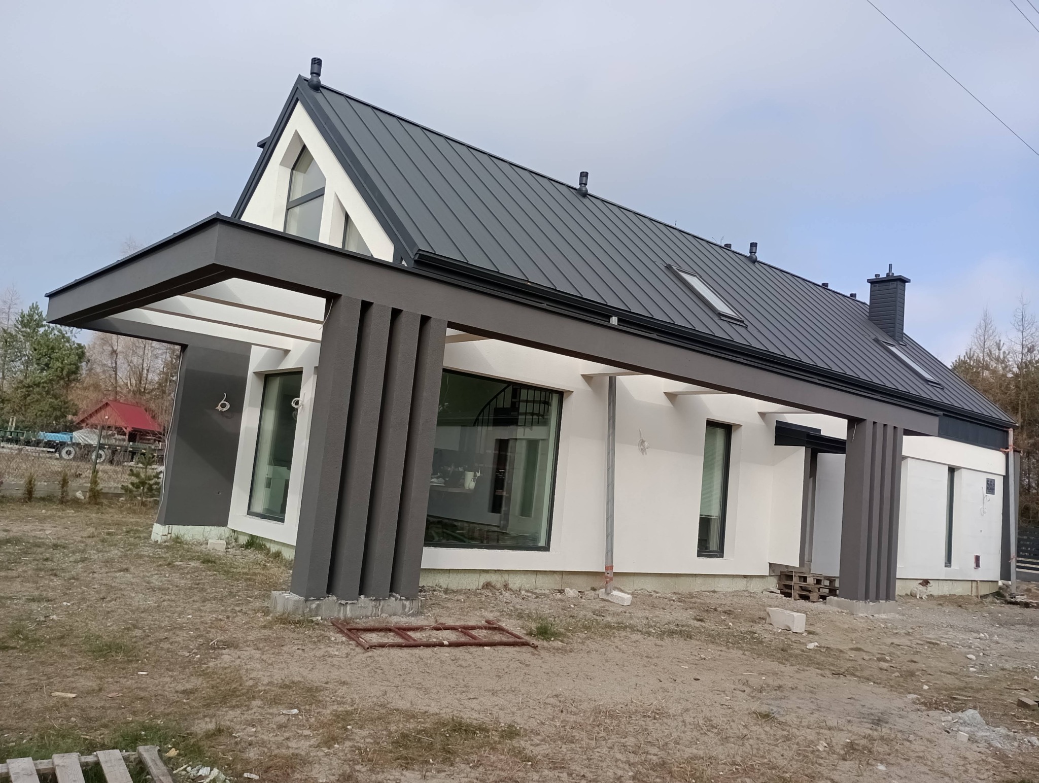 Nowoczesny dom jednorodzinny z szarą, metalową elewacją dachu i białymi ścianami. Widoczne duże okna i elementy konstrukcyjne w kolorze szarym. Teren wokół domu w trakcie prac wykończeniowych.