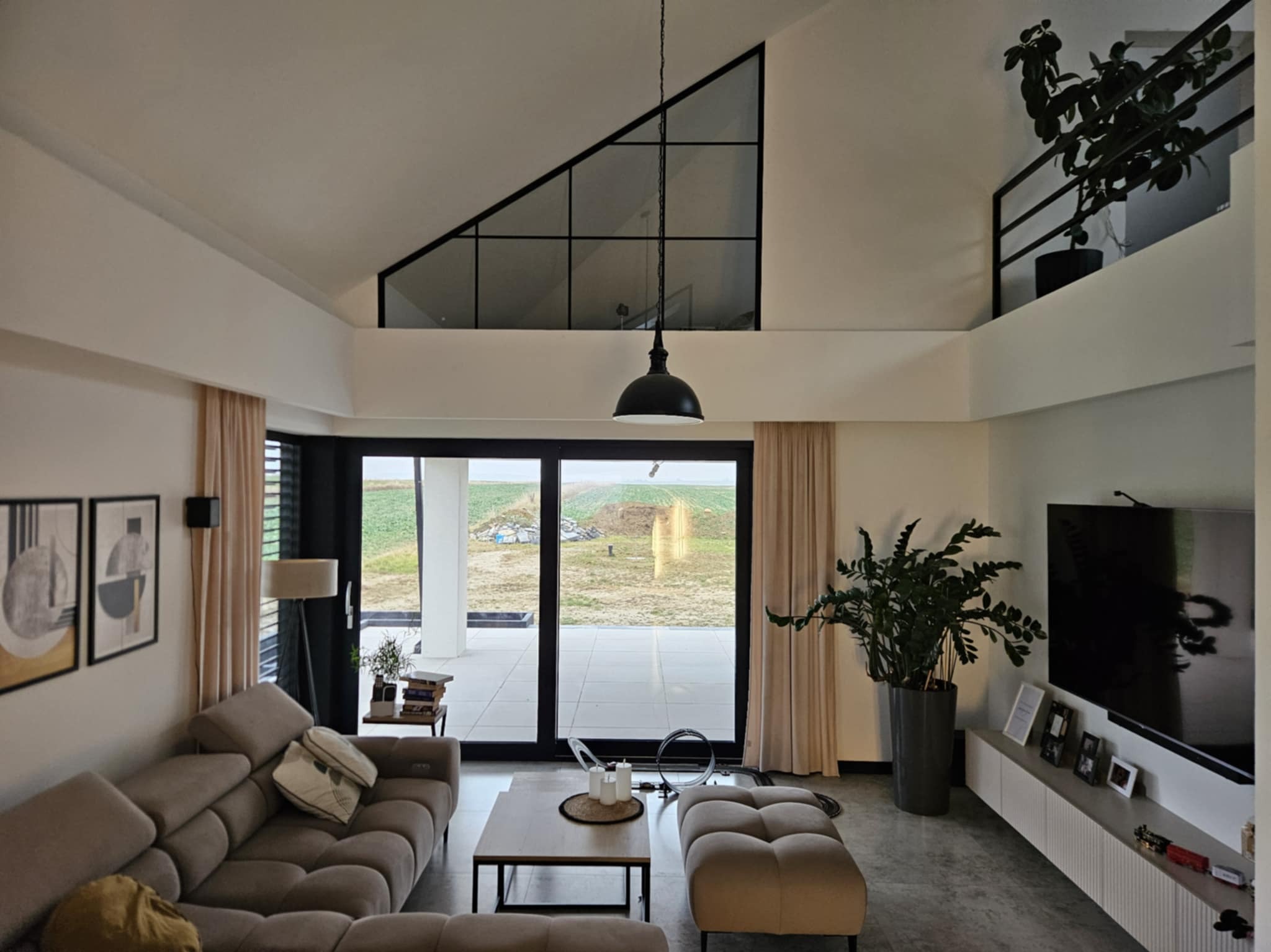 Jasne wnętrze salonu z dużym oknem na ogród. Minimalistyczny design, beżowa sofa, drewniany stolik kawowy, wysokie okno trójkątne. Rośliny doniczkowe dodają naturalnego akcentu.