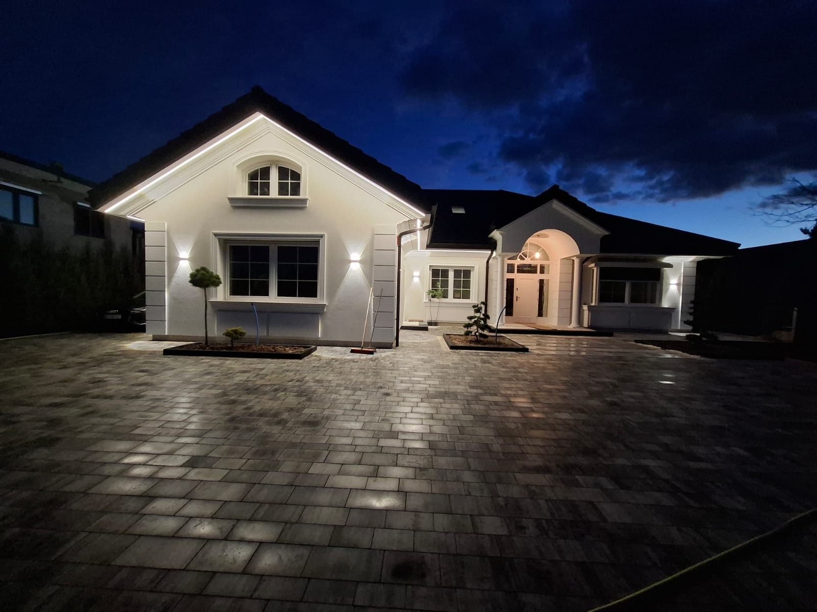 Elegancki dom jednorodzinny z oświetloną elewacją w Miączynie, sfotografowany nocą. Widoczny podjazd z kostki brukowej i starannie wykończona fasada budynku.