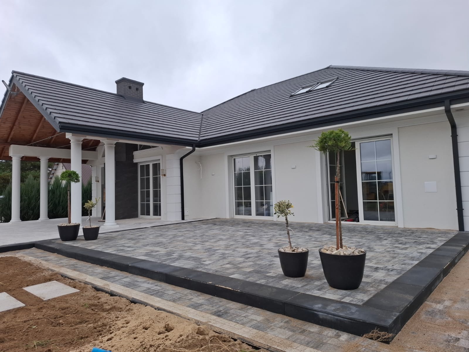 Nowoczesny dom z tarasem wyłożonym kostką brukową w odcieniach szarości. Biała elewacja, duże okna przesuwne i kolumnada dodają elegancji. Uwagę zwracają donice z drzewkami.