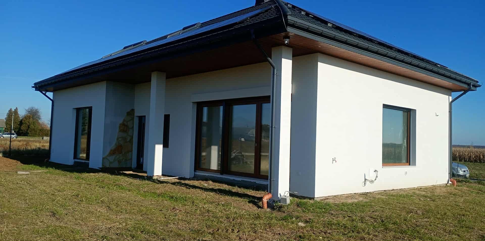 Nowoczesny dom jednorodzinny z białą elewacją, brązowymi oknami i panelami słonecznymi na dachu, otoczony zielonym trawnikiem w słoneczny dzień. Widok z boku.