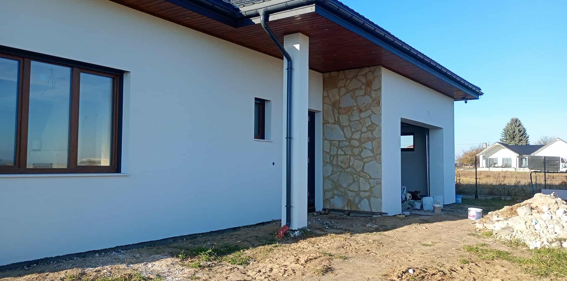 Wykończona elewacja domu jednorodzinnego z fragmentem ściany wyłożonym kamieniem dekoracyjnym. Widoczne okna z brązowymi ramami i elementy orynnowania.