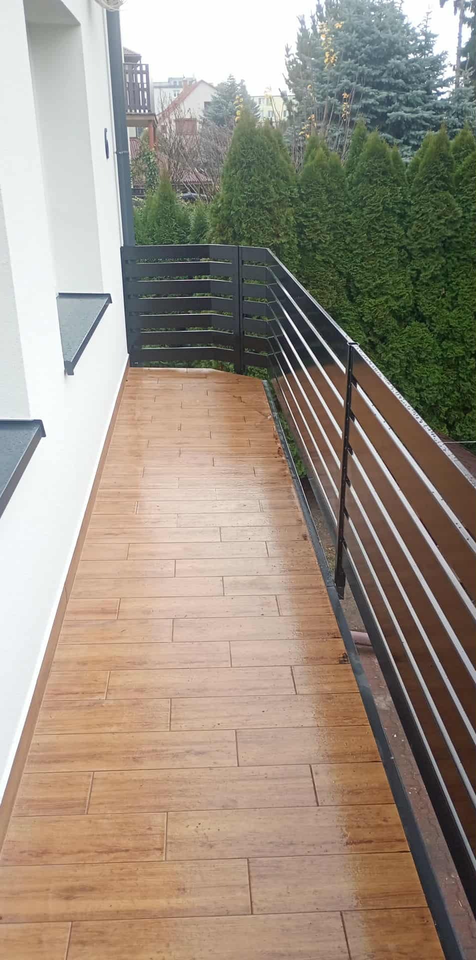 Balkon z drewnopodobną terakotą i nowoczesną balustradą z poziomych listew. Widok na zieleń. Ujęcie z góry, podkreślające wzór płytek i minimalistyczny design.