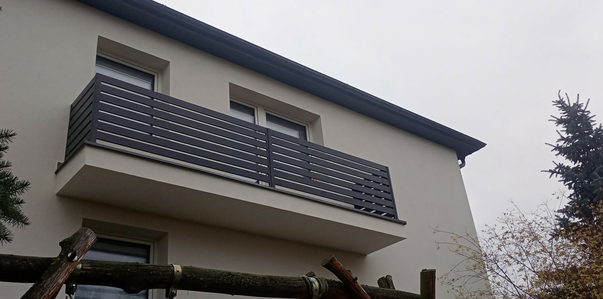 Elewacja budynku z balkonem o nowoczesnej, poziomej balustradzie w kolorze grafitowym. Widoczne okna i fragmenty drzew. Pochmurne niebo w tle.