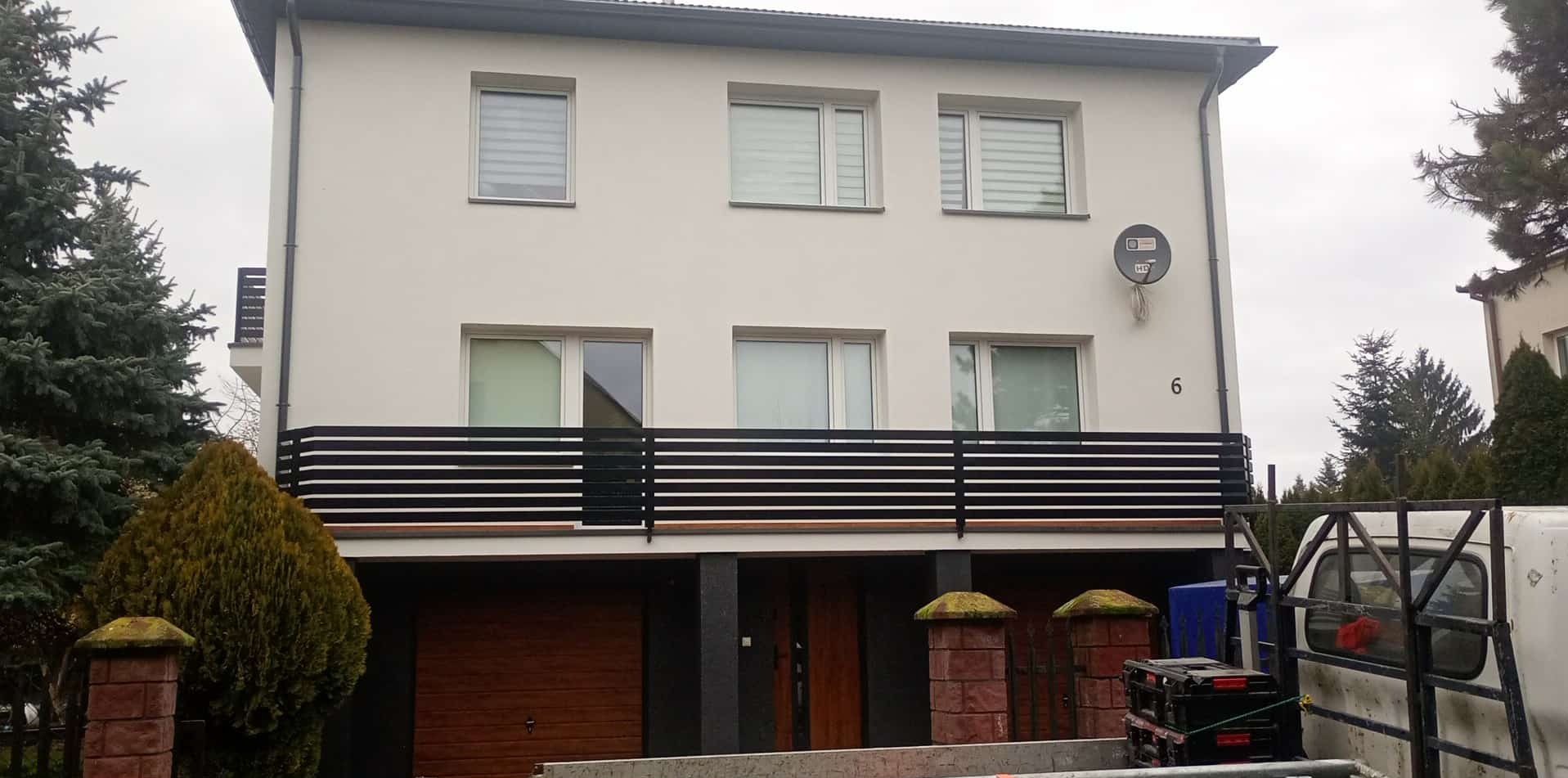 Dwukondygnacyjny dom z jasną elewacją, czarną balustradą balkonu i numerem 6. Widok z poziomu ulicy, częściowo zasłonięty przez roślinność i samochód dostawczy.