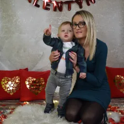 Kobieta w zielonej sukience trzymająca małego chłopca wskazującego palcem w górę, czerwone poduszki w serca, dekoracja 'VALENTYNKI' i płatki róż na podłodze.