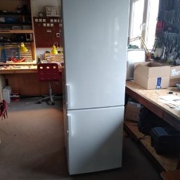 jarkinss Serwis Usługi - Biała lodówka marki Gorenje stoi w warsztacie, na tle stołu z narzędziami i urządzeniami pomiarowymi.