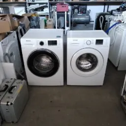 Dwie białe pralki Samsung stoją obok siebie w magazynie pełnym części zamiennych i sprzętu AGD, widoczne metalowe skrzynki narzędziowe i regały z różnymi elementami.