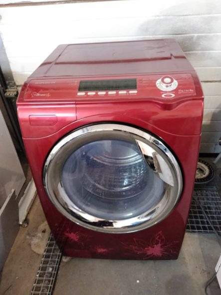 Czerwona pralka automatyczna z chromowanym wykończeniem drzwiczek i wyświetlaczem, widoczne motywy kwiatowe na obudowie, ustawiona na betonowej podłodze w garażu.