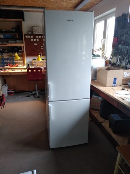 Biała lodówka marki Gorenje stoi w warsztacie, na tle stołu z narzędziami i urządzeniami pomiarowymi.