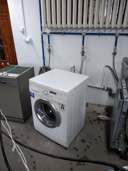 Biała pralka automatyczna z widocznym panelem sterowania i oznaczeniem klasy energetycznej A, stojąca obok srebrnej zmywarki w pomieszczeniu z grzejnikiem żeberkowym i instalacją hydrauliczną...