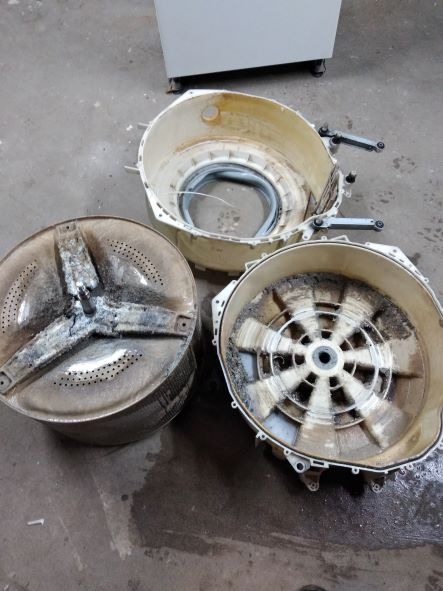 Rozłożona na części brudna pralka automatyczna: bęben z widocznymi osadami, obudowa z gumowym uszczelnieniem, oraz tylna część bębna z zanieczyszczeniami, na tle betonowej podłogi.