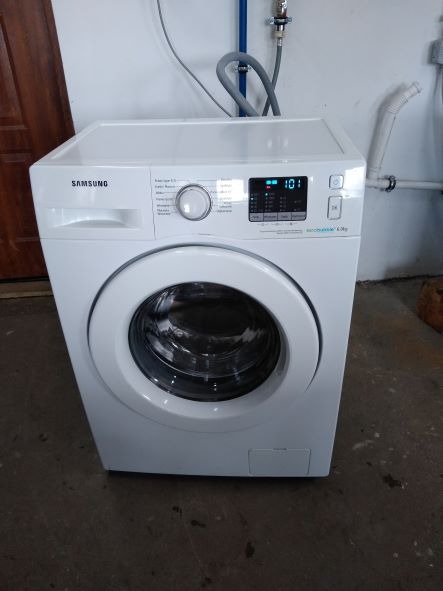 Biała pralka Samsung Eco Bubble 6.0 kg w pomieszczeniu z widocznymi rurami instalacji wodnej.