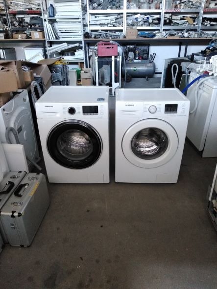 Dwie białe pralki Samsung stoją obok siebie w magazynie pełnym części zamiennych i sprzętu AGD, widoczne metalowe skrzynki narzędziowe i regały z różnymi elementami.