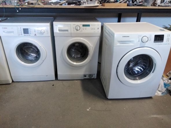 Trzy białe pralki różnych marek (Bosch, inna marka, Samsung) ustawione obok siebie na betonowej podłodze w warsztacie, z półką pełną kabli i części nad nimi.