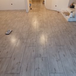 Slawex a-z - Ułożona podłoga z płytek imitujących drewno w odcieniach szarości, widoczna gąbka do fugowania, ściany przygotowane do malowania, widok na korytarz.