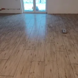 Ułożona podłoga z paneli imitujących drewno, widok z góry na ułożone panele, w tle okno z widocznym zabawką na zewnątrz, na podłodze narzędzie do układania paneli.