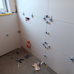 Slawex a-z - Ściana w trakcie układania płytek, widoczne krzyżyki poziomujące, przewody instalacyjne i narzędzia na podłodze.