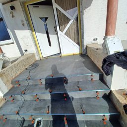 Slawex a-z - Układanie ciemnoszarych płytek na schodach wejściowych do domu, z użyciem systemu poziomowania płytek (klipsy i kliny), widoczny kabel zasilający i cień osoby robiącej zdjęcie.