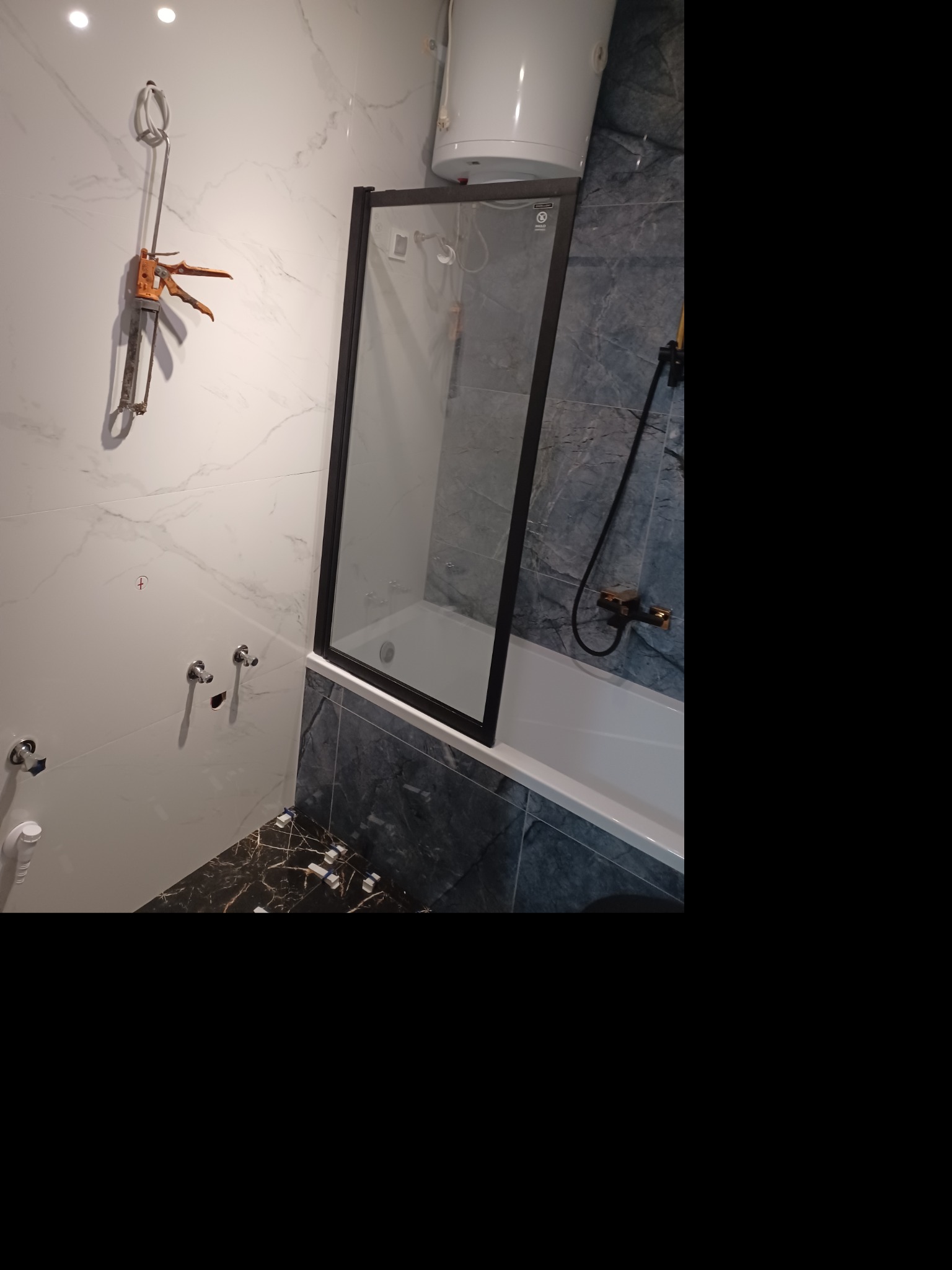 Nowoczesna łazienka w trakcie wykańczania: białe marmurowe płytki, czarna armatura, kabina prysznicowa z czarną ramą i narzędzie do silikonu zamocowane na ścianie.