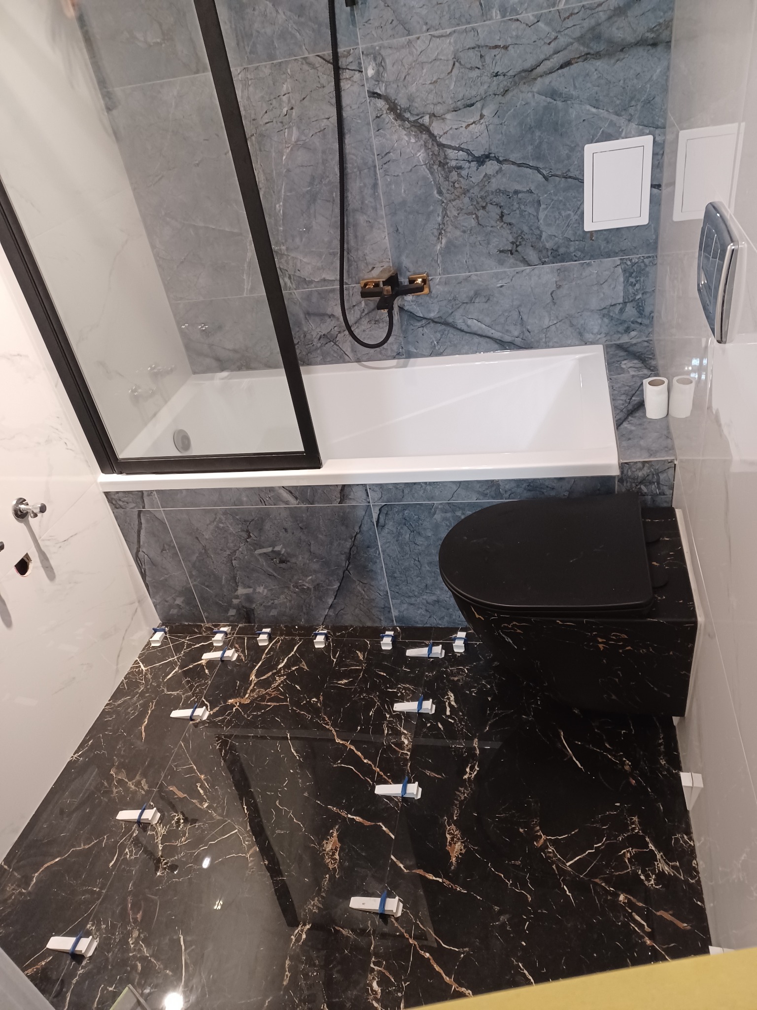 Wnętrze łazienki w trakcie prac wykończeniowych: czarna toaleta i płytki podłogowe imitujące marmur z białymi krzyżykami dystansowymi, ściany wyłożone szarymi płytkami z marmurowym wzorem, biała...