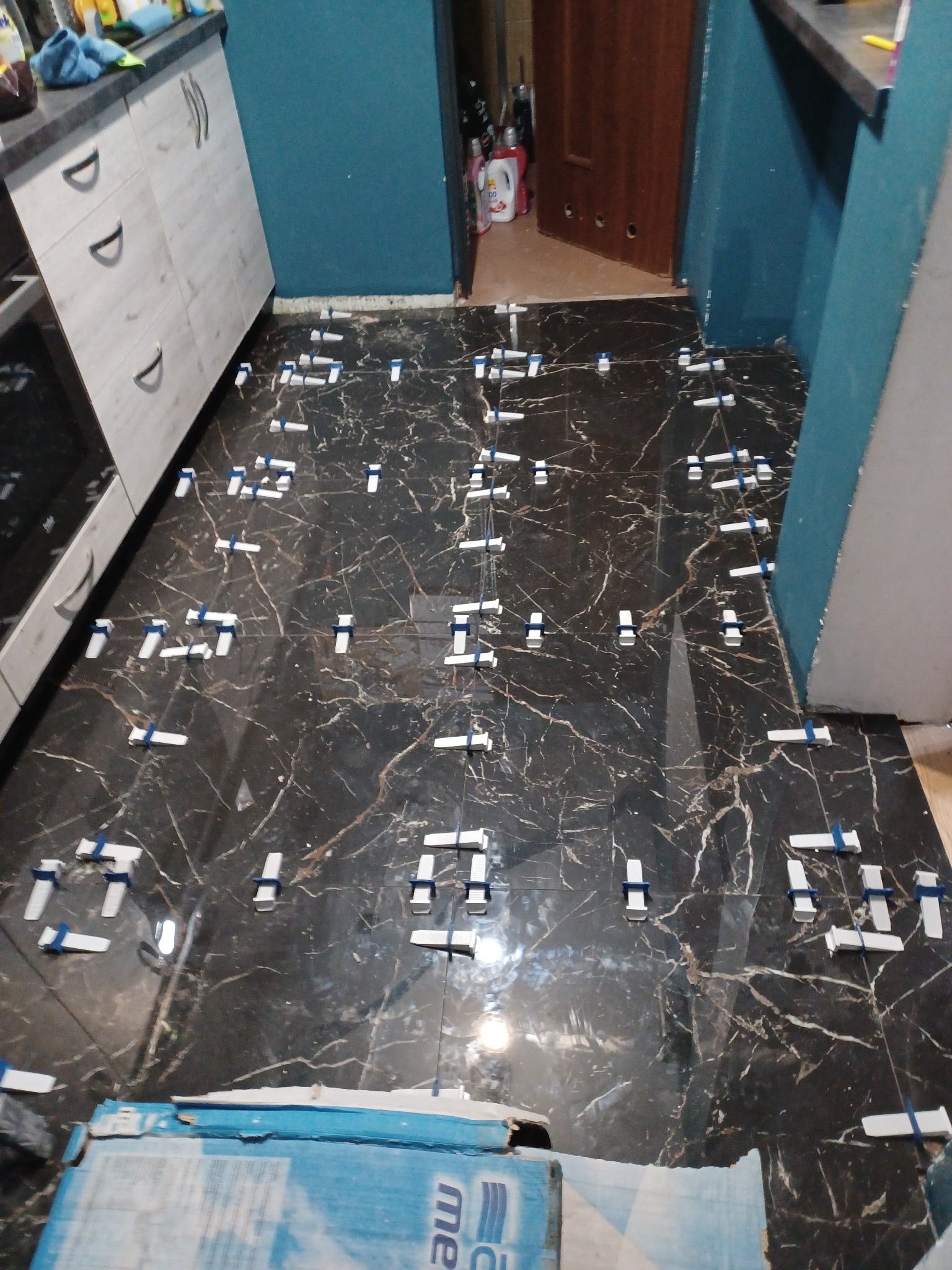 Czarne płytki imitujące marmur układane na podłodze w kuchni, z użyciem białych krzyżyków dystansowych z niebieskimi wstawkami, widoczne fragmenty szafek kuchennych i ściany pomalowanej na niebiesko.