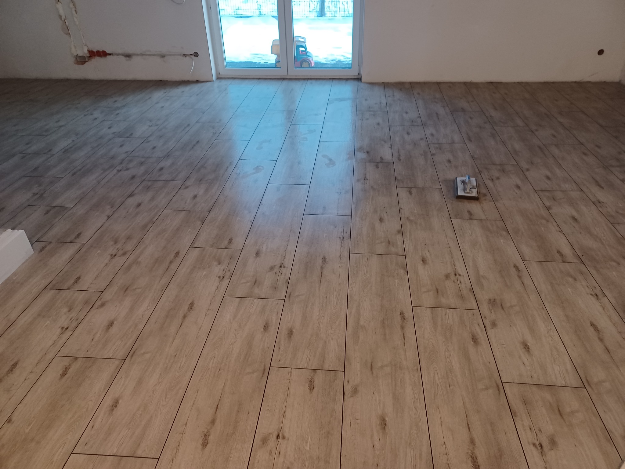 Ułożona podłoga z paneli imitujących drewno, widok z góry na ułożone panele, w tle okno z widocznym zabawką na zewnątrz, na podłodze narzędzie do układania paneli.