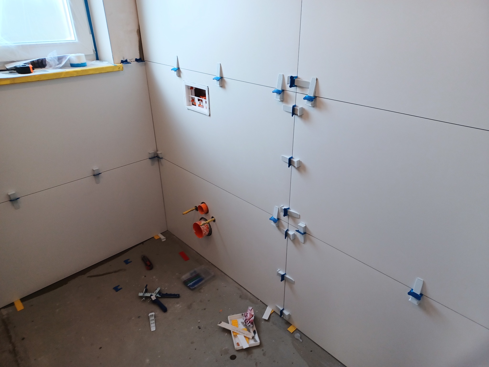 Ściana w trakcie układania płytek, widoczne krzyżyki poziomujące, przewody instalacyjne i narzędzia na podłodze.