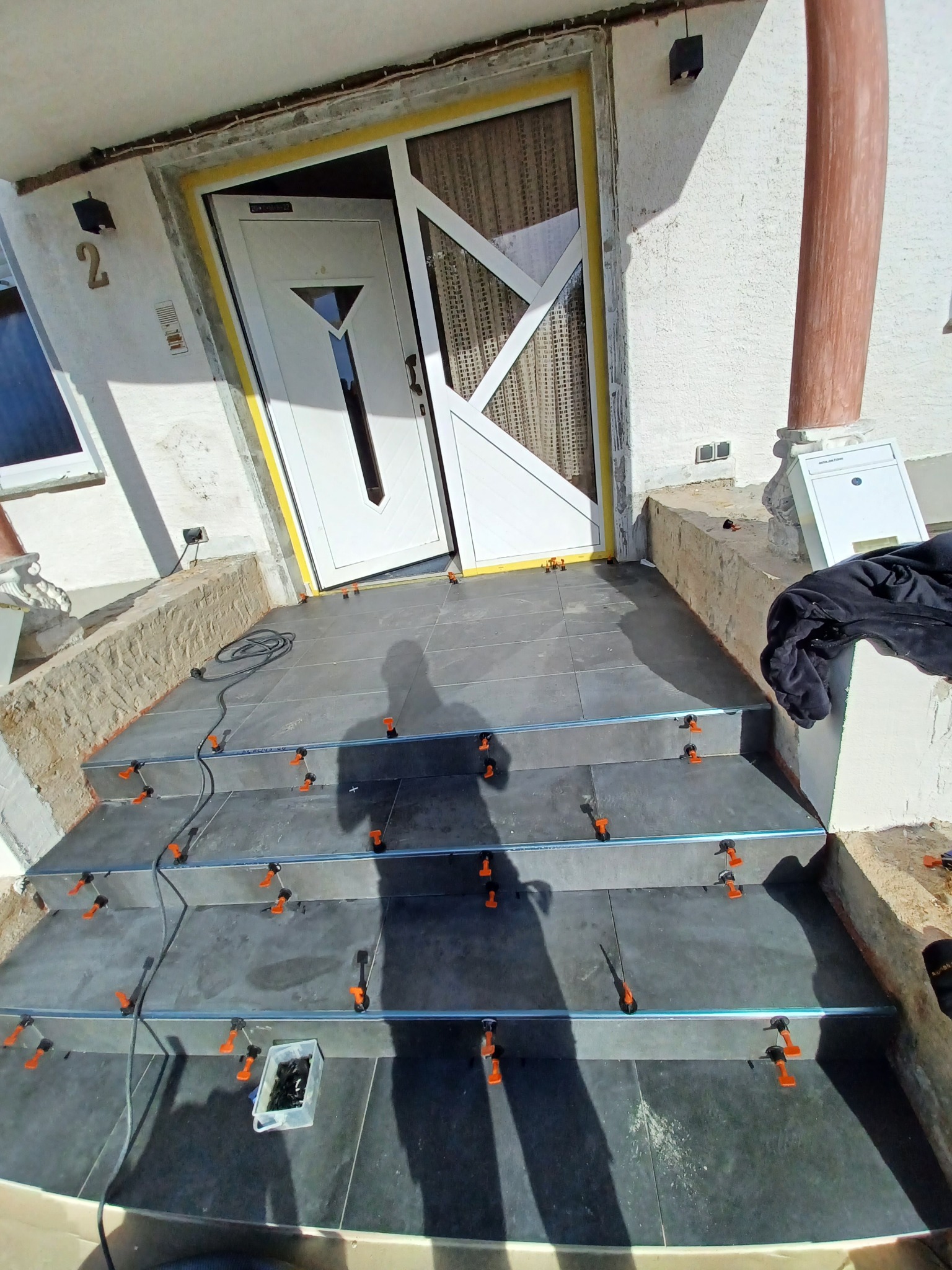 Układanie ciemnoszarych płytek na schodach wejściowych do domu, z użyciem systemu poziomowania płytek (klipsy i kliny), widoczny kabel zasilający i cień osoby robiącej zdjęcie.