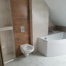 Wnętrze łazienki w trakcie prac wykończeniowych: biała wanna, podwieszana muszla klozetowa z chromowanym przyciskiem, płytki imitujące drewno na ścianie, szare płytki podłogowe, wiertarko-wkrętarka...