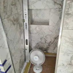 Poziomica oparta o ścianę z marmurowymi płytkami w trakcie prac we Wrocławiu, z widoczną wnęką i wentylatorem na podłodze.