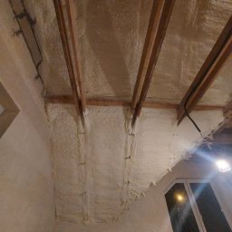 SOLID PARTNER Michał Szemfeld - Poddasze po natrysku izolacji pianką PUR, widoczne krokwie i elementy konstrukcyjne dachu, ściany również pokryte pianką, okno w tle.