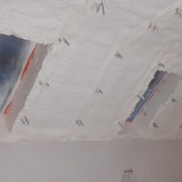 SOLID PARTNER Michał Szemfeld - Poddasze ocieplone białą pianką PUR z widocznymi łatami i elementami montażowymi, fragment ściany w dolnej części kadru.