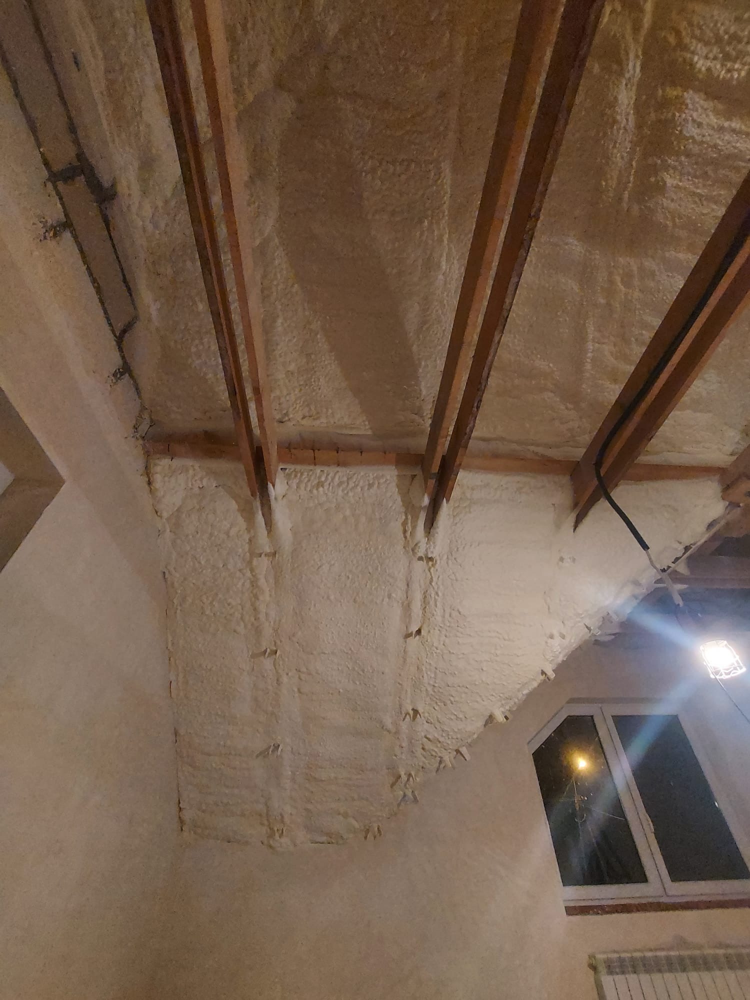 Poddasze po natrysku izolacji pianką PUR, widoczne krokwie i elementy konstrukcyjne dachu, ściany również pokryte pianką, okno w tle.
