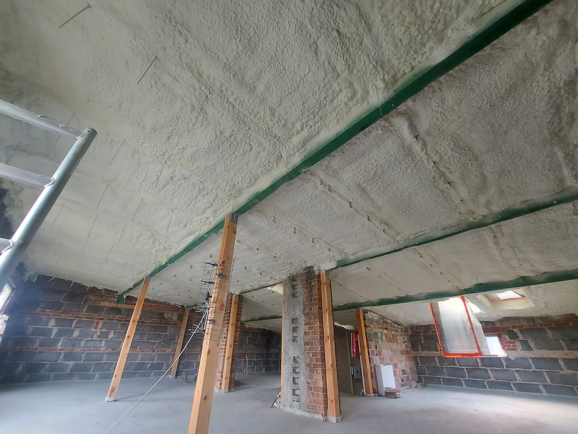 Poddasze z odsłoniętą cegłą, ocieplone białą pianką PUR, widoczne drewniane słupy podtrzymujące konstrukcję dachu i drabina oparta o ścianę.