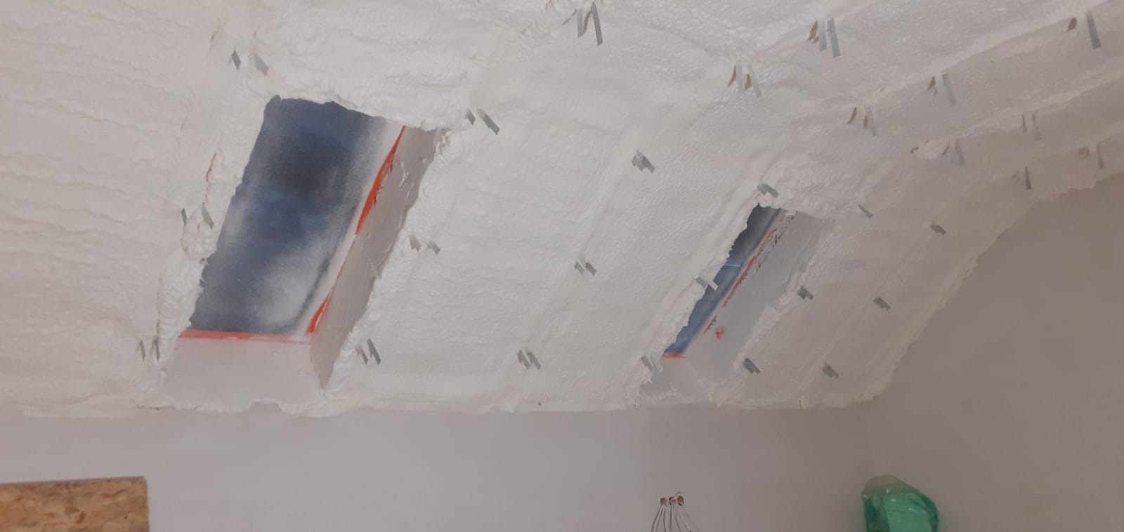 Poddasze ocieplone białą pianką PUR z widocznymi łatami i elementami montażowymi, fragment ściany w dolnej części kadru.