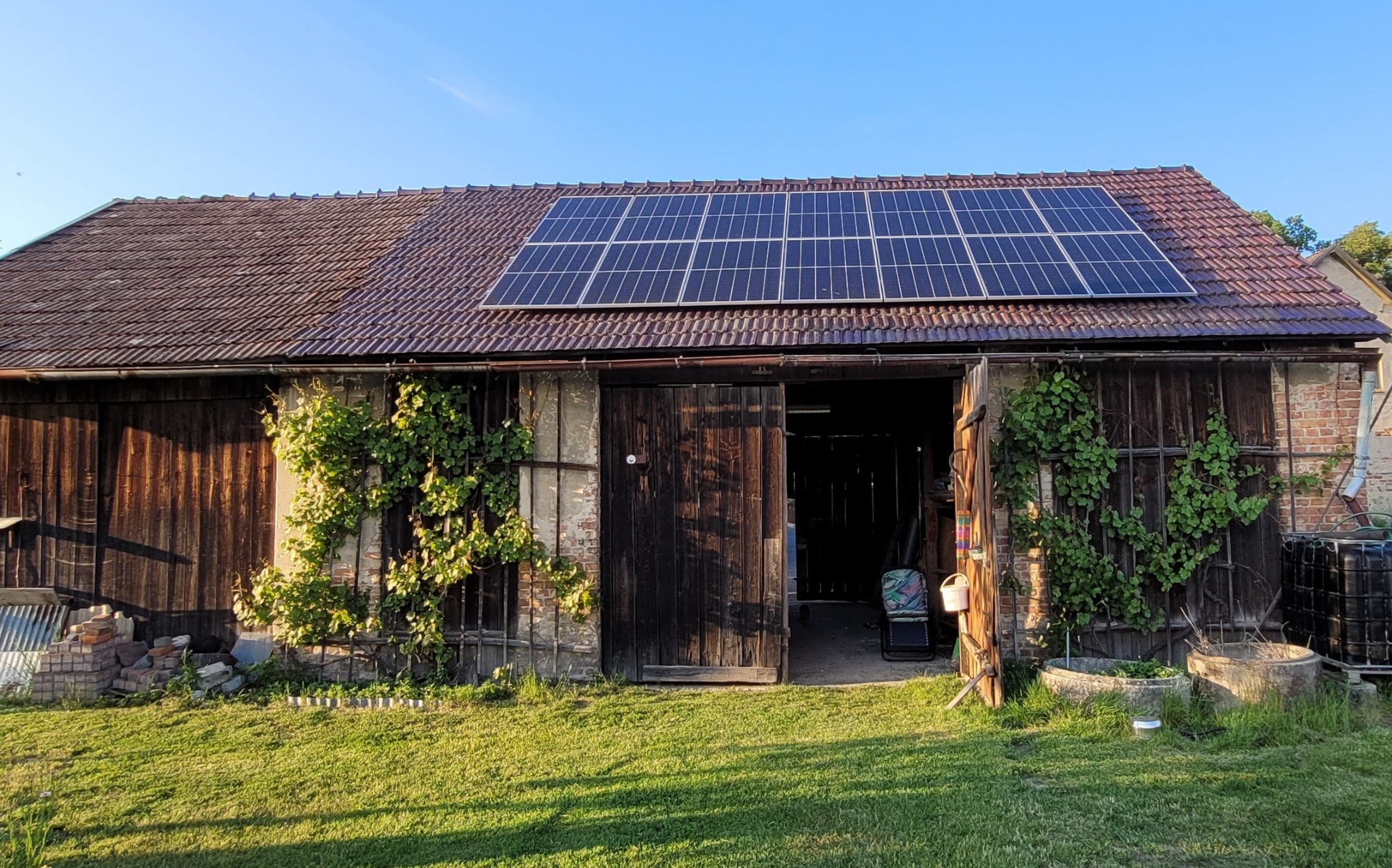 Wiejski budynek gospodarczy z zainstalowanymi panelami słonecznymi na dachu z czerwonej dachówki, z otwartymi drewnianymi wrotami i pnącą roślinnością na ścianach.